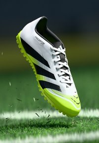 Chaussure de football blanche et jaune néon avec des rayures noires, dessus texturé et semelle à crampons ; en l'air au-dessus d'une pelouse verte.