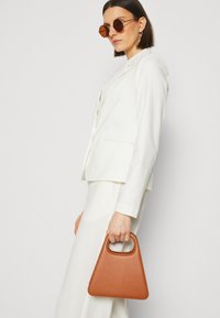 Weißer Blazer über einem weißen Rock, hält eine dreieckige braune Lederhandtasche. Runde braune Sonnenbrillen und minimalistische Designdetails sind sichtbar.