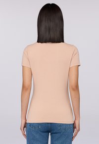 Koszulka z krótkim rękawem, dopasowana, wykonana z miękkiego beżowego materiału, z okrągłym dekoltem. Tył ma gładką fakturę i brak widocznych oznaczeń.