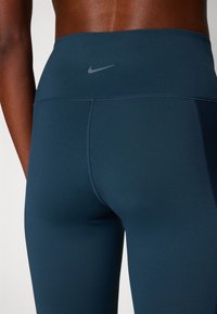 Magas derékú, sötétkék, elasztikus anyagból készült leggings, karcsú fazonban, varrás nélküli kialakítással, a derékpánton pedig egy kicsi szürke Nike logó található.