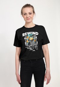 Mladá žena v černém tri�čku s barevným robotem a texty včetně "REWIND" a "ASC CRAWLER", stojící na neutrálním pozadí.
