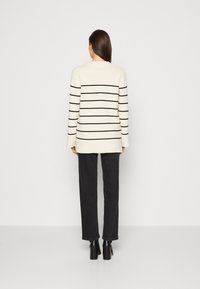 Suéter crema a rayas con líneas horizontales negras, mangas largas, abertura leve en el dobladillo, combinado con pantalones negros de pierna ancha y botas con tacón.