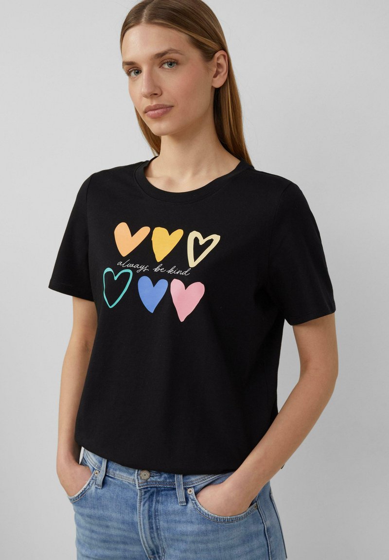 T-shirt en coton noir à manches courtes, avec des graphismes de cœurs multicolores et le texte "soyez toujours gentil" affiché en évidence sur le devant.