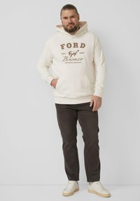 Kremowa bluza z kapturem z brązowym napisem "FORD" i logo; kieszeń kangurka; w połączeniu z ciemnobrązowymi spodniami i białymi trampkami. Gładka tkanina, luźny krój.