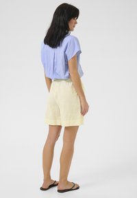 Light blue short-sleeve blouse with back pleat, paired with yellow elastic-waist shorts featuring a cuffed hem, worn with black flip-flops.
