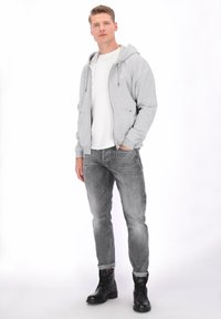 Sweat à capuche gris avec fermeture éclair, en mélange de coton, porté sur un T-shirt blanc, associé à un jean gris délavé et des bottes noires à lacets.