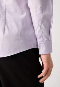 Cămașă butonată de culoare violet deschis, cu o textură fină, având un mansetă cu un singur buton și un tiv rotunjit, asortată cu pantaloni închisi la culoare.