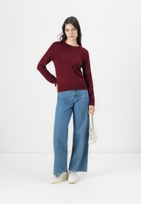 Bordo pleten sweater v kombinaciji s svetlo modrimi širokimi hlačami in belimi supergami, drži majhno belo torbico.