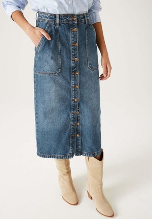 Jupe en denim à coupe droite, couleur bleue, fermeture à boutons à l'avant, deux poches latérales et détails de couture. Associée à des bottes beiges jusqu'aux genoux.
