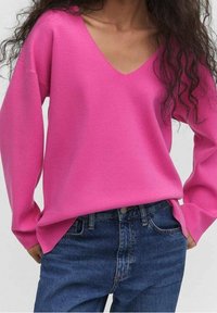 Rosa Strickpullover mit tiefem V-Ausschnitt, langen Ärmeln und gerippten Bündchen, kombiniert mit blauen Jeans mit vorderer Knopfleiste und Taschen.