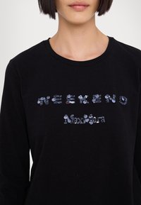 Donna che indossa un top nero a maniche lunghe con "Weekend Max Mara" in paillettes luccicanti sul petto.