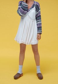 Weißes Kleid mit einem gefalteten Rock und Spitzenakzenten, kombiniert mit einem mehrfarbigen, gemusterten Langarmoberteil. Braune Holzclogs und graue Socken.