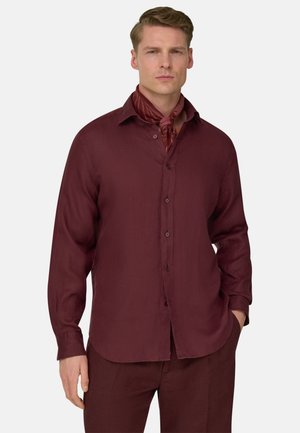 Homme portant une chemise rouge foncé à boutons et un pantalon assorti, avec une écharpe rouge à motifs nouée autour du cou, debout avec une main dans la poche.