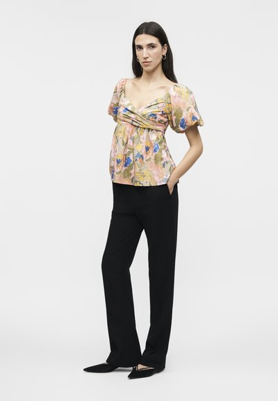 Γυναίκα στέκεται με το ένα χέρι στην τσέπη, φορώντας μπλούζα με φουσκωτά μανίκια και λουλούδια, με πιέτες μπροστά, μαύρο παντελόνι ίσιας γραμμής και μαύρα μυτερά παπούτσια.