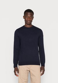 Marineblå strikkegenser med teksturert mønster, rund halslinning og ribbestrikkede mansjetter og linning. Bærer den med lys beige bukser.