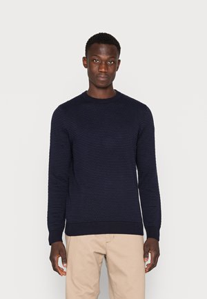 Marineblå strikkegenser med teksturert mønster, rund halslinning og ribbestrikkede mansjetter og linning. Bærer den med lys beige bukser.
