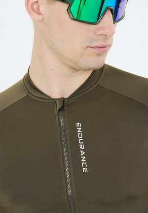 Nærbillede af en person iført en olivengrøn sportstrøje med lynlås og teksten "ENDURANCE" samt reflekterende sportsbriller med grønligt skær.