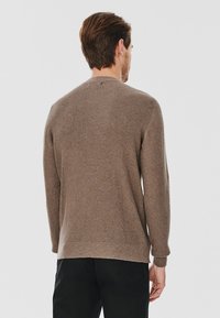 Brązowy sweter robiony na drutach z teksturowanym wzorem, okrągłym dekoltem i długimi rękawami. Dół jest nieco dłuższy z tyłu, co zapewnia luźny fason.