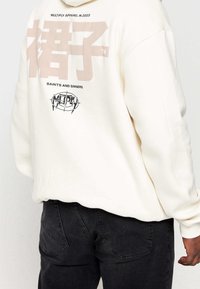 Felpa crema con grandi grafiche beige sul retro, comprese scritte e un logo circolare, abbinata a jeans neri. Design semplice e casual.