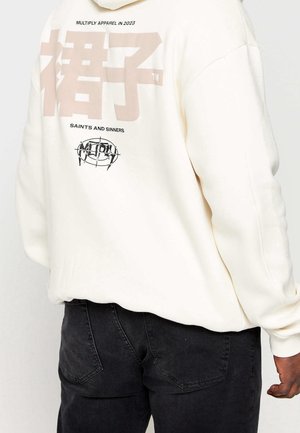 Cream sweatshirt with large beige graphics on the back, including text and a circular logo, paired with black jeans. Simple, casual design.