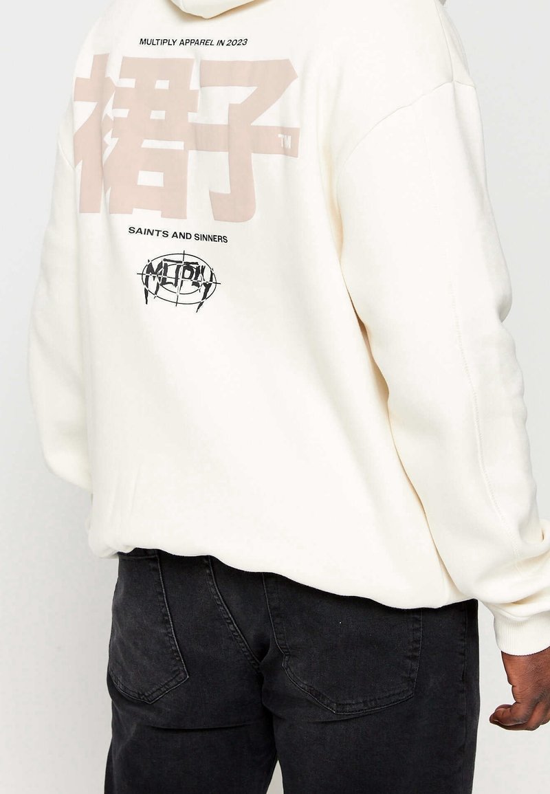 Felpa crema con grandi grafiche beige sul retro, comprese scritte e un logo circolare, abbinata a jeans neri. Design semplice e casual.