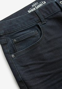 Blugi din denim închis, cu o vedere detaliată a brâului, care prezintă detalii cusute, un buzunar cu închidere tip clichet și o etichetă de marcă.