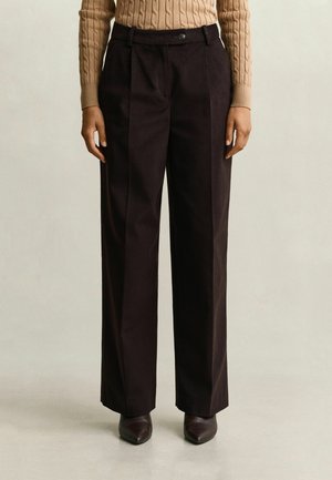 Femme portant un pantalon large taille haute marron foncé avec un pli sur le devant, associé à un pull beige en maille torsadée et des chaussures foncées.