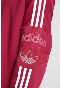 Bordeaux atletisk jacka med tre vita ränder på ärmarna, meshdetaljer och en vit Adidas-logotyp på ärmen.