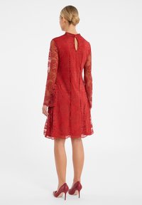 Robe en dentelle rouge avec de longues manches transparentes, détail de dos en forme de serrure, ourlet festonné et silhouette ajustée, associée à des talons hauts rouges.