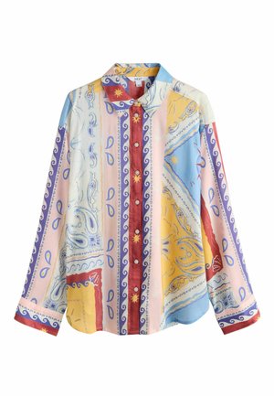 Nő egy kőfalnak támaszkodva, mintás felsőt és fehér csíkos nadrágot viselve, zöld cserepes növények mellett áll szabadban.