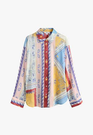 Moteris atsiremianti į akmeninę sieną, dėvinti raštuotą marškinių švarką ir baltas dryžuotas kelnes, stovinti šalia žalių vazoninių augalų lauke.