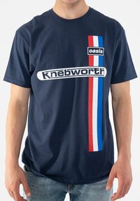 Navy blue cotton t-shirt featuring white "oasis" and "Knebworth" text. Red, white, and blue vertical stripes accent the left side.