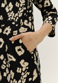 Zwarte jurk met een beige dierenprint, met een zichtbare zijzak. De stof heeft een gladde textuur en een ontspannen pasvorm.