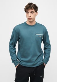 Νεαρός άνδρας που φοράει πετρόλ μακρυμάνικο πουκάμισο με τσέπη στο στήθος με φερμουάρ και μαύρο παντελόνι με λογότυπα μάρκας, στέκεται με τα χέρια στις τσέπες.