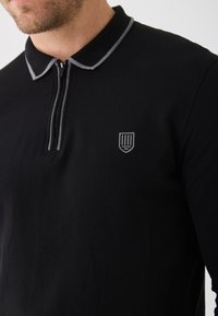 Svart pikétröja av texturerad tyg, med dragkedja i halsen, grå krage och en liten broderad sköldlogotyp på bröstet.