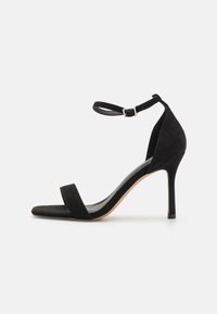 Musta mokkanahkainen korkokenkä, jossa on nilkkaremmli, avoin varvas ja hoikka stiletto-korko. Ominaisuuksiin kuuluvat minimalistinen muotoilu ja pehmeä rakenne.