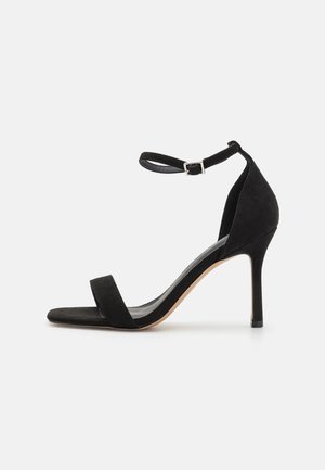 Sandalo con tacco alto in suede nero, dotato di cinturino alla caviglia, punta aperta e sottile tacco a stiletto. Presenta un design minimalista e una texture morbida.