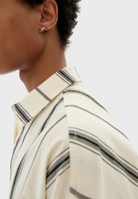Camisa a rayas en crema y negro. Presenta un diseño con cuello y un cierre frontal con botones. Tejido de algodón suave, con detalles de costura visibles.