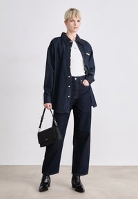 Mørk denimskjorte med knapper, hvit T-skjorte under, høytlivede denimbukser og en svart håndveske. Modellen har på seg svarte høye støvler.