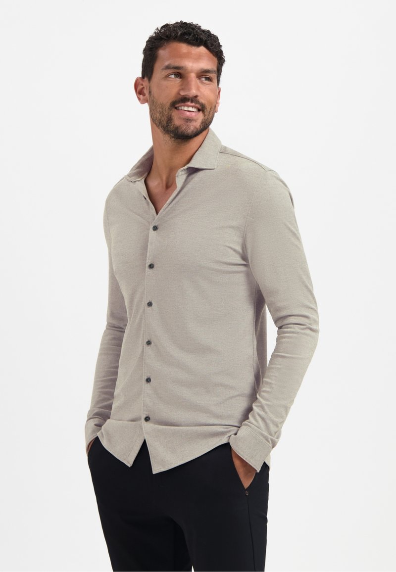 Lichtgrijs langemouwen shirt met een subtiele textuur, een puntige kraag en donkere knopen. Draagt met een zwarte broek, handen in de zakken.