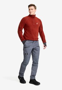 Man i rostfärgad fleece med kvartslång dragkedja, grå cargobyxor med lappar och svarta vandringsskor, stående med händerna i fickorna.