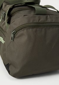 Grønn stoff duffelbag med teksturert overflate, med flere glidelåslommer og robuste håndtak. Kompakt rektangulær form.