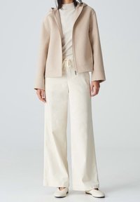 Femme portant une veste beige zippée, un pantalon large taille haute crème clair avec cordon de serrage, et des chaussures plates crème sur fond gris.