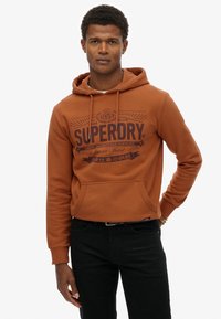 Brązowa bluza z kapturem z przednią kieszenią, z logo "SUPERDRY" w ciemnym brązie. Miękki materiał, o luźnym kroju, noszona z czarnymi spodniami.