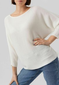 Weißer gerippter Strickpullover mit weitem Ausschnitt und Dreiviertelärmeln, kombiniert mit blauen Jeans. Weiche Textur mit lockerer Passform.