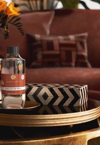 Klar glassflaske med Suede Vanilla hjemmeparfume refill med svart hette, plassert på en dekorativ svart og hvit geometrisk eske på et gullfat.