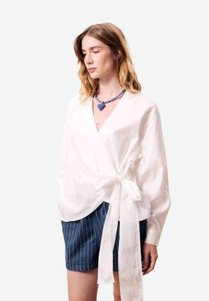 Vrouw met lichtbruin haar, gekleed in een witte wikkelblouse die in de taille is gestrikt, blauwe shorts met fijne strepen en een blauwe ketting met een hartvormige hanger van kralen.