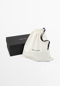 Sac en coton avec cordon de serrage de couleur crème avec le logo "Massimo Dutti", à côté d'une boîte rectangulaire noire à fini mat.