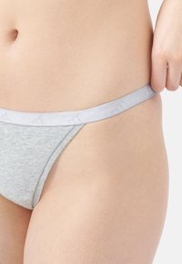 Detailní záběr osoby upravující pas světle šedých bavlněných spodních kalhot na boku proti jednoduchému pozadí.