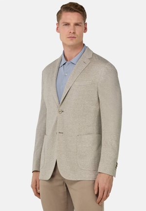 Blazer en chevrons beige avec deux poches avant, fermeture à un bouton et revers crantés, porté sur un polo bleu clair. Texture lisse.
